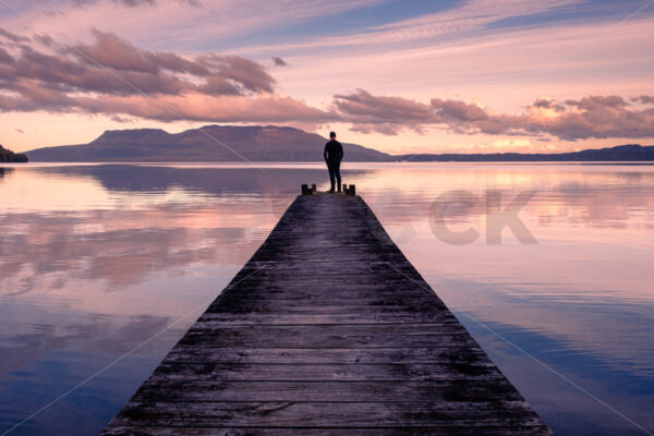 A man standing on a Lake Tarawera jetty at sunset with Mount Tarawera in the distance, Rotorua, Bay of Plenty, New Zealand - SCP Stock