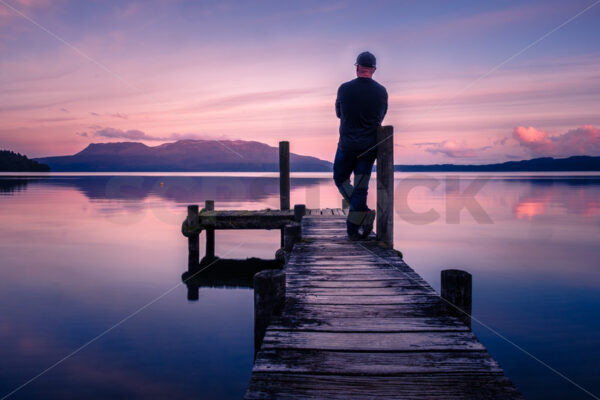 A man standing on a Lake Tarawera jetty at sunset with Mount Tarawera in the distance, Rotorua, Bay of Plenty, New Zealand - SCP Stock