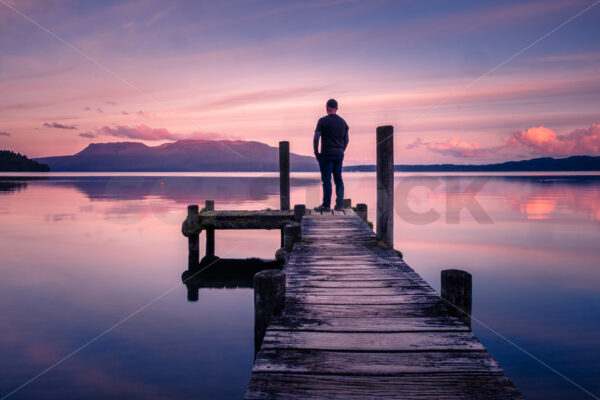 A man standing on a Lake Tarawera jetty at sunset with Mount Tarawera in the distance, Rotorua, Bay of Plenty, New Zealand - SCP Stock