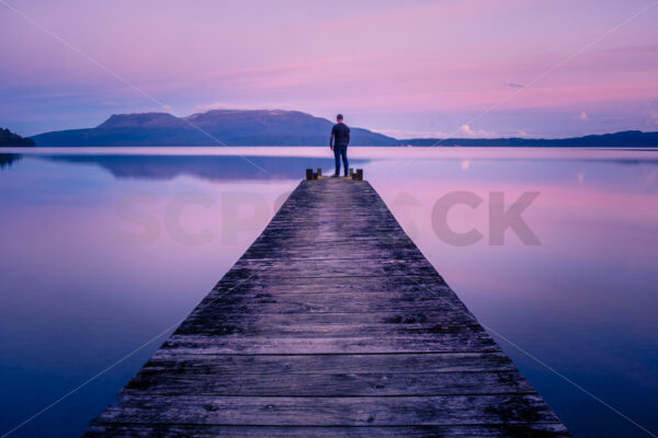 A man standing on a Lake Tarawera jetty at sunset with Mount Tarawera in the distance, Rotorua, Bay of Plenty, New Zealand - SCP Stock