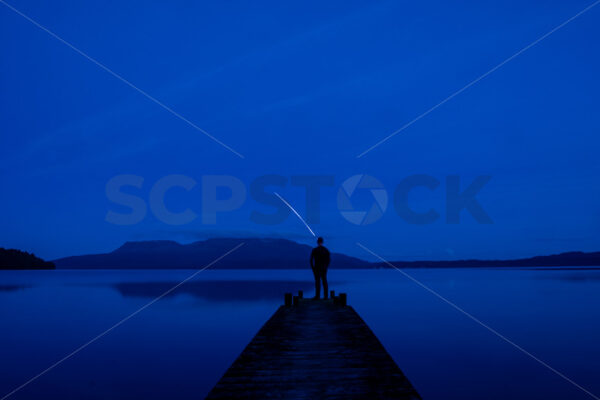 Rocket Man – A man standing on a Lake Tarawera jetty at night watching a Rocket Lab launch, Rotorua, Bay of Plenty, New Zealand (composite image) - SCP Stock